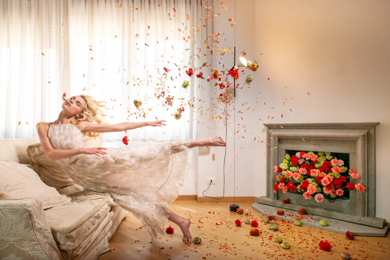 Stefano Delìa, La Scala di Mohs, scatto n.5 – donna in abito chiaro sospesa in movimento con petali e fiori che riempiono la stanza.