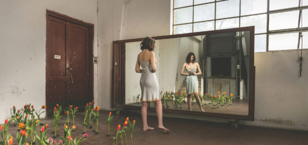 Stefano Delìa, fotografia autoriale dal progetto La Scala di Mohs, scatto n.2 con donna davanti a uno specchio e tulipani in ambiente industriale