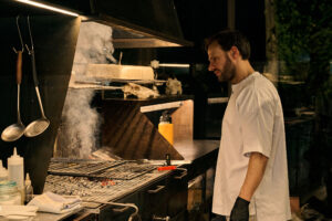 Lo chef Edoardo Tilli osserva la griglia ardente in cucina, con brace e utensili da lavoro