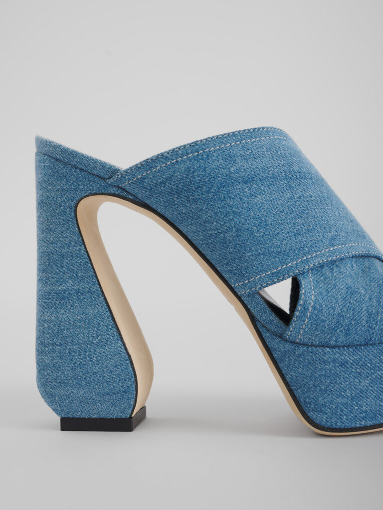 Mule in denim con tacco scultoreo, fotografata sullo stesso fondo grigio e con la stessa luce del resto del catalogo